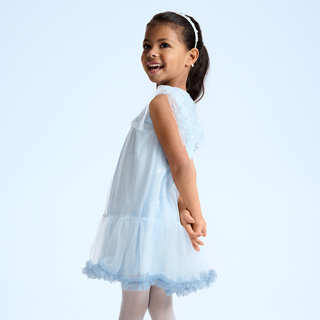 A young girl posing in a light, flowing dress standing with hands clasped behind her back. The image highlights the elegance and delicate design of the outfit. banner