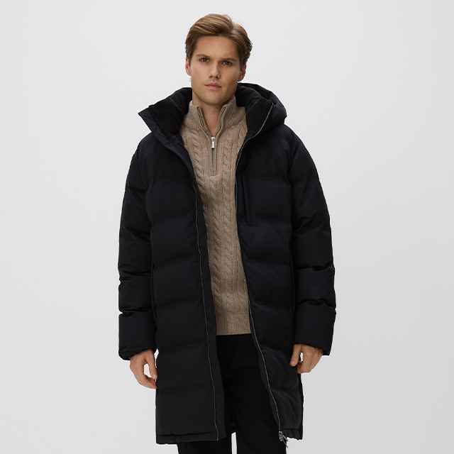 A person wearing a long, black quilted winter coat with a hood, paired with a beige knitted sweater featuring a quarter-zip design and dark pants. The outfit appears warm and suitable for colder weather conditions. banner