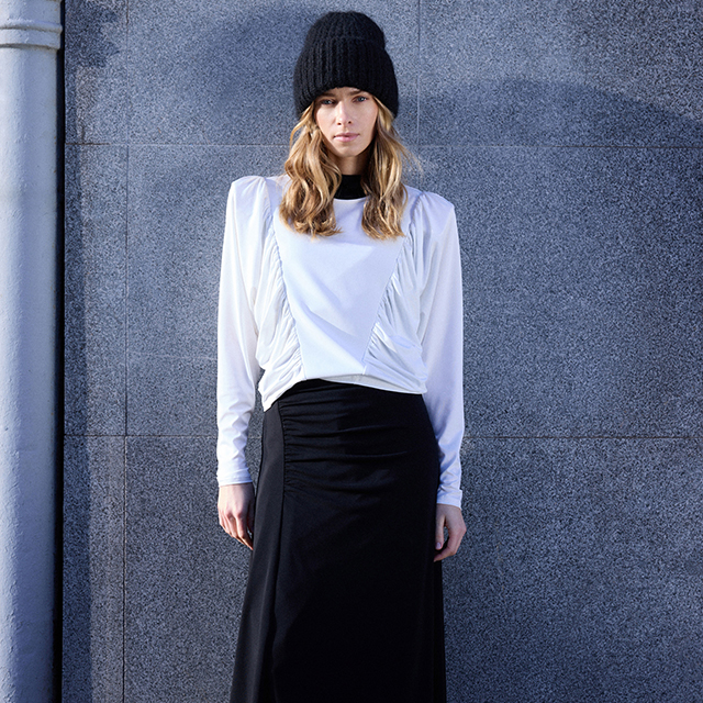 A person wearing a white long-sleeve top, a black skirt, and a black knit hat standing in front of a gray textured wall.