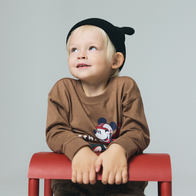 A child is sitting cross-legged on the floor, wearing a festive knitted sweater with a snowflake pattern, grey pants, and brown shoes. The scene evokes a cozy and relaxed atmosphere. banner