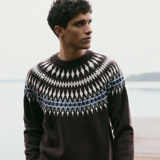 A person wearing a knitted sweater with a geometric pattern featuring white shapes and blue details. The background includes a calm body of water and a soft, cloudy sky. The sweater design draws attention with its intricate style and cozy look, perfect for colder weather. banner