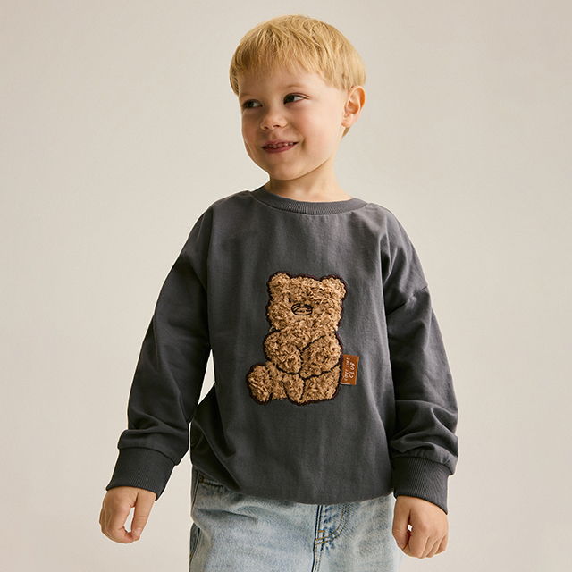 A child is wearing a white T-shirt with a Messi graphic design and black pants. The child holds a black hoodie in one hand and poses confidently against a neutral background.