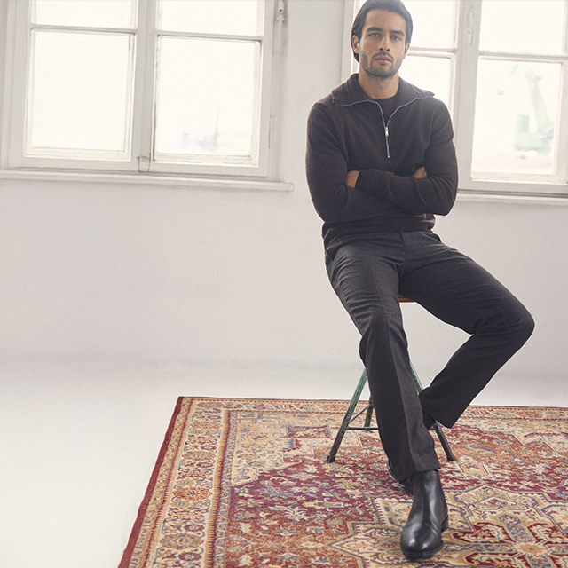 A person sits casually on a stool placed on a patterned rug in a minimalist room. Bright natural light streams through two large windows behind them, creating a serene atmosphere. The pose and setting suggest a relaxed moment indoors. banner