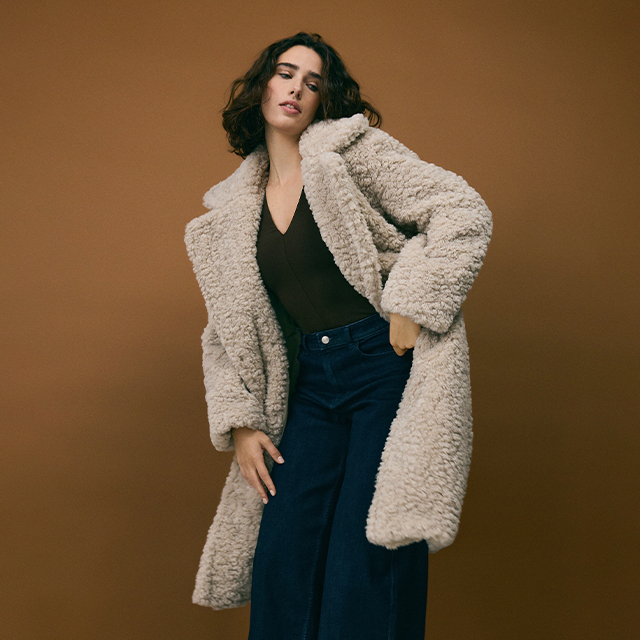 A person wearing a sparkling sleeveless gown paired with a large fur coat draped over their shoulders, standing against a neutral background. banner