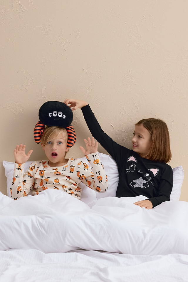 Two children sitting on a bed covered with white bedding. One child wears pajamas with fox prints and holds up their hands, while the other child, dressed in cat-themed pajamas, playfully places a stuffed spider toy with striped legs on the first child’s head. banner