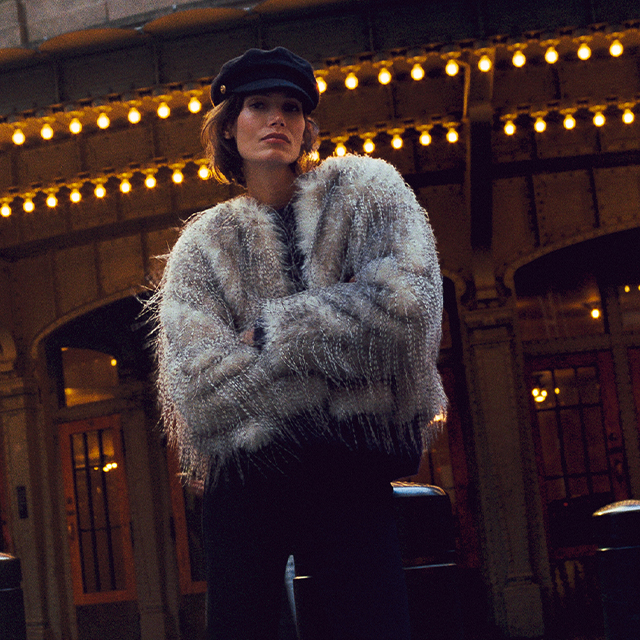 A person is standing outdoors at night beneath string lights, wearing a soft, textured jacket and a cap, with arms crossed. The scene has a warm urban atmosphere, with illuminated windows and decorative lights in the background. banner