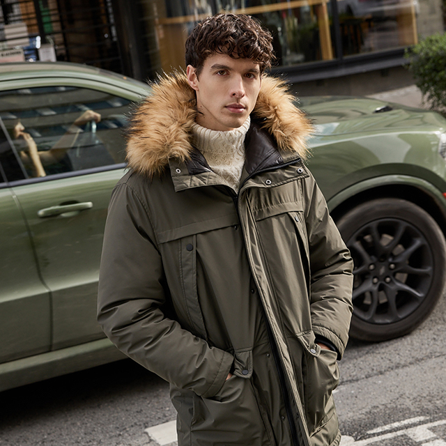 A person is wearing a winter jacket with a fur-lined hood, standing outdoors near a green parked car on an urban street. The scene suggests casual winter attire suitable for cold weather. The background includes storefronts and urban elements. banner