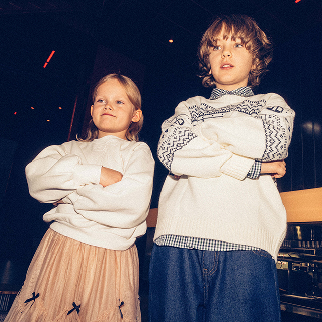 Two children stand confidently with arms crossed, wearing winter clothing: one in a sweater with patterns and jeans, the other in a plain sweater and a skirt. They appear in a modern indoor setting, possibly a casual or event space. The atmosphere suggests a relaxed yet self-assured mood. banner