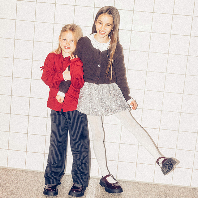 Two children are posing in stylish outfits against a tiled wall. One is wearing a red shirt and striped pants, while the other wears a white dress with tights and a cardigan. Both are showcasing playful and confident postures. banner