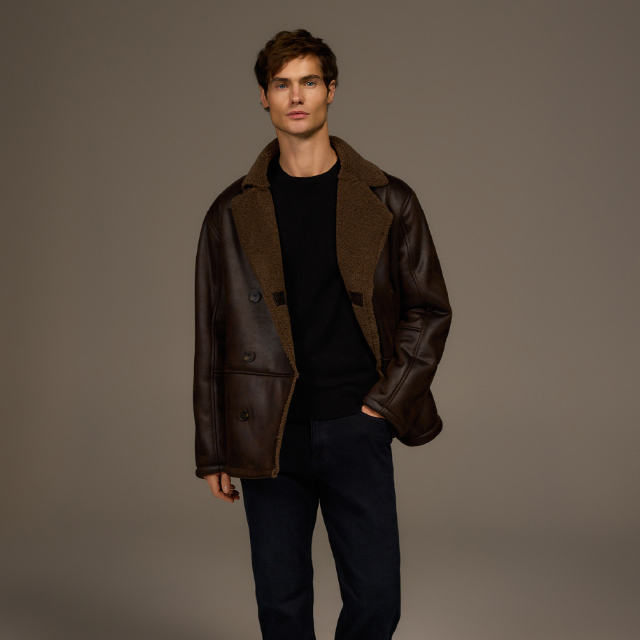 A person sits casually on a stool placed on a patterned rug in a minimalist room. Bright natural light streams through two large windows behind them, creating a serene atmosphere. The pose and setting suggest a relaxed moment indoors. banner