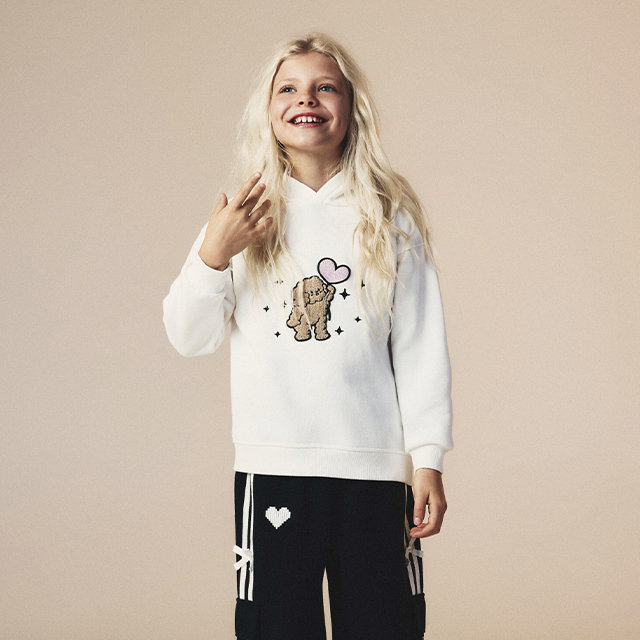 A person is wearing a white hoodie with a teddy bear graphic surrounded by stars and a heart. Paired with black pants featuring heart and stripe designs. Casual outfit suitable for everyday wear or lounging. Image emphasizes clothing details and design features. banner
