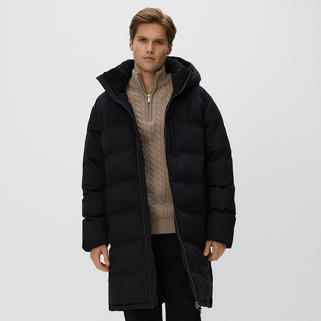 A person wearing a long, black quilted winter coat with a hood, paired with a beige knitted sweater featuring a quarter-zip design and dark pants. The outfit appears warm and suitable for colder weather conditions. banner