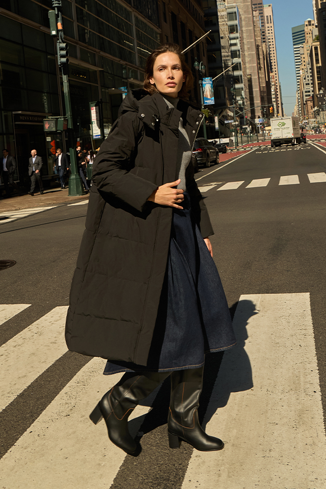 A person is walking across a crosswalk in a city surrounded by tall buildings. They are dressed in a long coat, a skirt, and high boots, carrying an object in their hand. The urban environment includes traffic lights, parked cars, and pedestrians in the background. banner