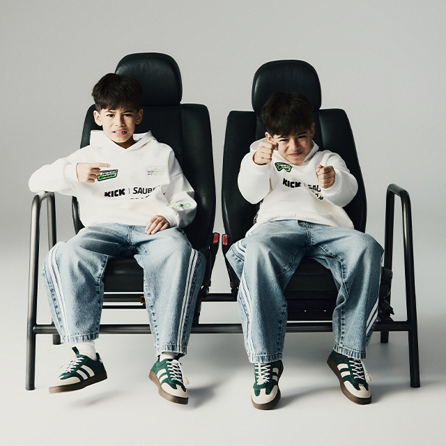 Two children are sitting on connected black chairs, wearing casual outfits with jeans, white hoodies, and green sneakers. Both are holding something in their hands and pointing forward, possibly pretending to aim or gesture. Their actions suggest playfulness or engagement in a game. banner