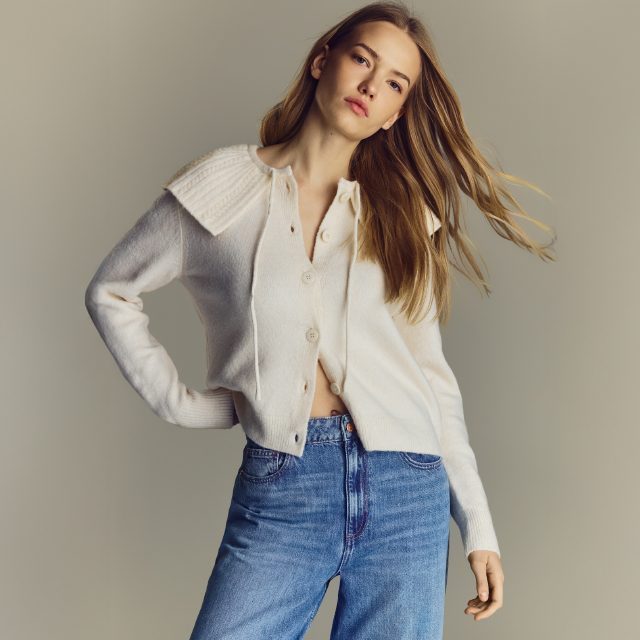 A person is wearing a white button-up cardigan with a wide collar and blue high-waisted jeans. The cardigan is slightly open, revealing a casual and relaxed style. Hair flows naturally, complementing the outfit. Suitable for everyday or casual occasions. banner