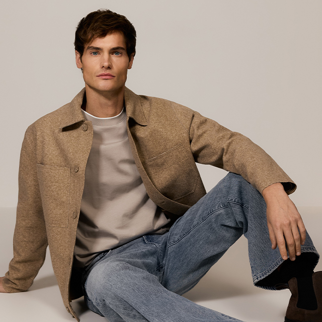 A person wearing a red sweater, black jeans, and a brown jacket with a visible shearling lining is standing in a neutral pose. They are also wearing a dark knitted hat, slightly adjusting the jacket with their hands.