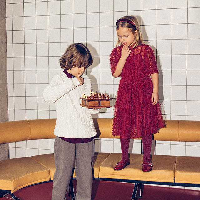 Two children are engaged in an activity. One is holding a small chessboard, examining it closely, while the other stands on a bench, observing. The setting includes a tiled wall and a yellow seating area, indicating an indoor environment. No facial details are visible. banner