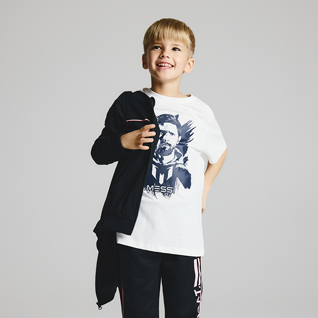 A child is wearing a white T-shirt with a Messi graphic design and black pants. The child holds a black hoodie in one hand and poses confidently against a neutral background. banner