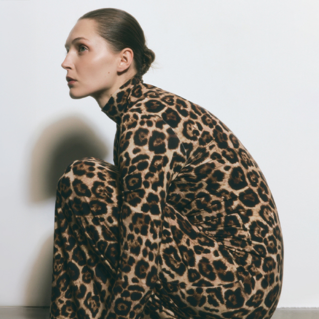 A person wearing a full-body, tight leopard print outfit is crouched down, positioned sideways, with a white background behind. The image emphasizes the patterned clothing style. banner