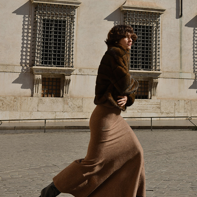 A person dressed in a long brown skirt and a fur jacket is walking outdoors near a historic building with barred windows and stone walls. The image captures a moment of motion, showcasing elegance and style in a cityscape setting. banner