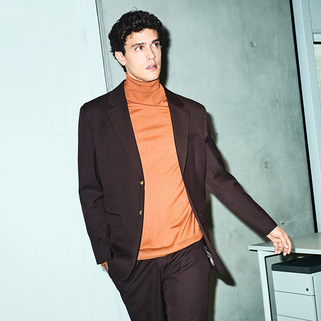 A person wearing a knitted sweater with a geometric pattern featuring white shapes and blue details. The background includes a calm body of water and a soft, cloudy sky. The sweater design draws attention with its intricate style and cozy look, perfect for colder weather. banner