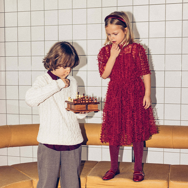 Two children are posing in stylish outfits against a tiled wall. One is wearing a red shirt and striped pants, while the other wears a white dress with tights and a cardigan. Both are showcasing playful and confident postures. banner