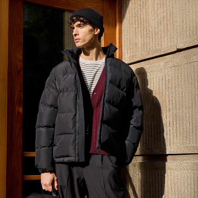 A person wearing a padded jacket, striped shirt, burgundy cardigan, and black pants stands outside near a wooden door and a stone wall, holding a bag in one hand. Sunlight illuminates part of the scene, casting shadows on the wall. banner