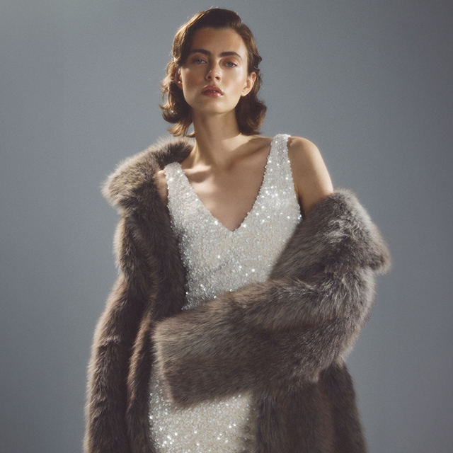 A person wearing a sparkling sleeveless gown paired with a large fur coat draped over their shoulders, standing against a neutral background. banner