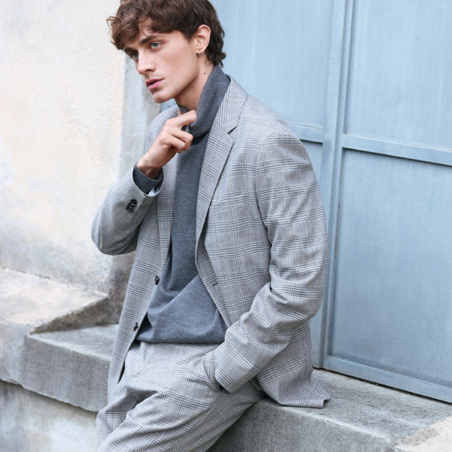 A person wearing a long, black quilted winter coat with a hood, paired with a beige knitted sweater featuring a quarter-zip design and dark pants. The outfit appears warm and suitable for colder weather conditions. banner