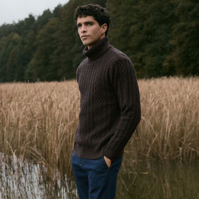 Person wearing a dark cable-knit turtleneck sweater and blue pants, standing near a calm lake surrounded by tall dry grass and dense green forest in the background. banner