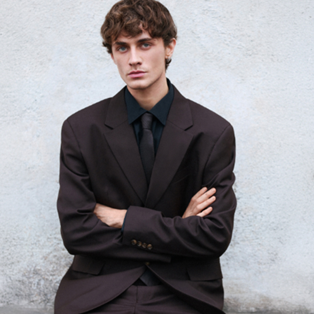 A person wearing a dark suit and tie sits with arms crossed against a plain background. banner