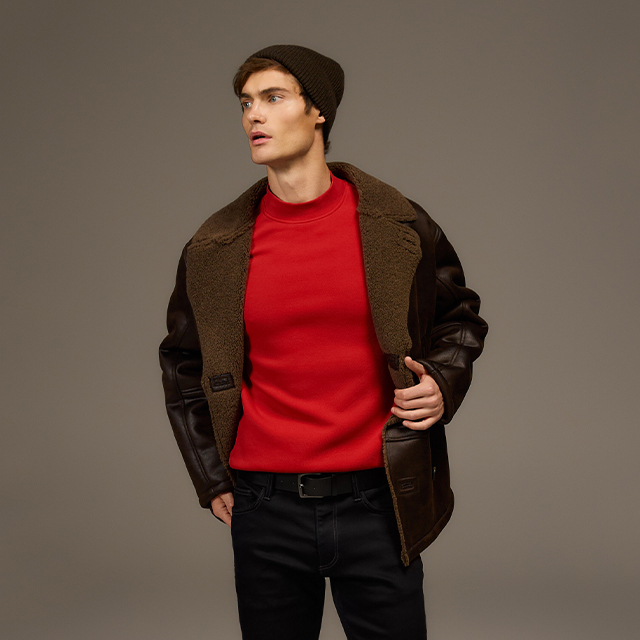 A person wearing a red sweater, black jeans, and a brown jacket with a visible shearling lining is standing in a neutral pose. They are also wearing a dark knitted hat, slightly adjusting the jacket with their hands.