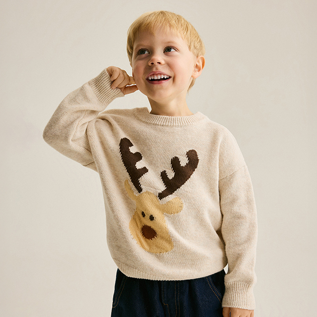 A child is wearing a white T-shirt with a Messi graphic design and black pants. The child holds a black hoodie in one hand and poses confidently against a neutral background.