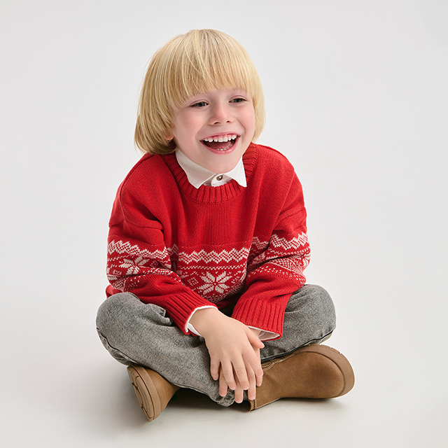 A child is sitting cross-legged on the floor, wearing a festive knitted sweater with a snowflake pattern, grey pants, and brown shoes. The scene evokes a cozy and relaxed atmosphere. banner