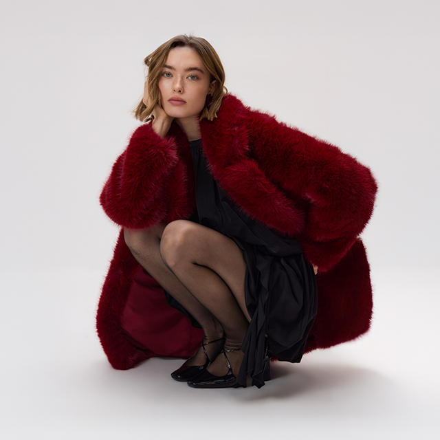 A person crouching while wearing a red faux fur coat over a black dress and black tights. The outfit appears elegant and stylish, portraying a bold fashion choice with a focus on texture and layered clothing. banner