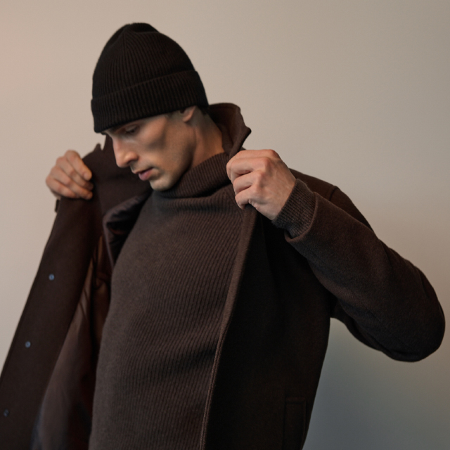 A person is adjusting their coat collar while wearing a knitted sweater and a beanie. The image captures the action of pulling up the coat for comfort or style. Perfect for showcasing winter clothing or outdoor apparel in a casual setting. banner